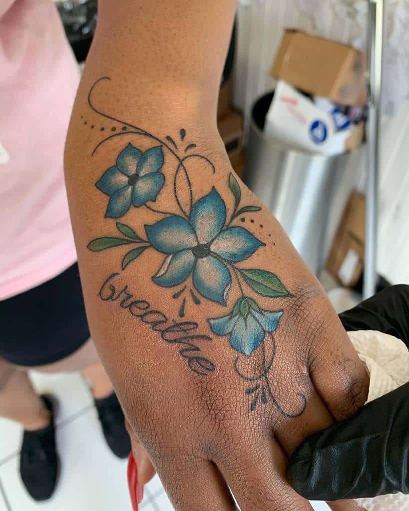 A stunning hand tattoo featuring blue flowers gracefully wraps around the word "breathe"