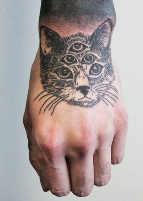 Hand tattoo of a cat with four eyes, inked in fine black linework with shading, blending into a dark gradient on the wrist.