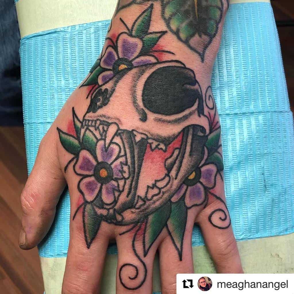 Hand tattoo of a roaring cat skull surrounded by purple flowers and greenery
