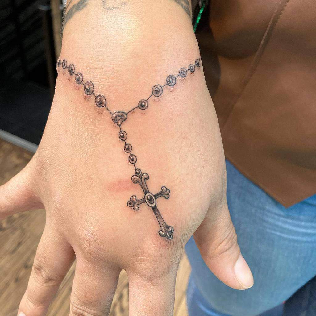 Tattoo of intricately detailed beads and a cross gracefully adorns the back of a hand, elegantly resembling a rosary