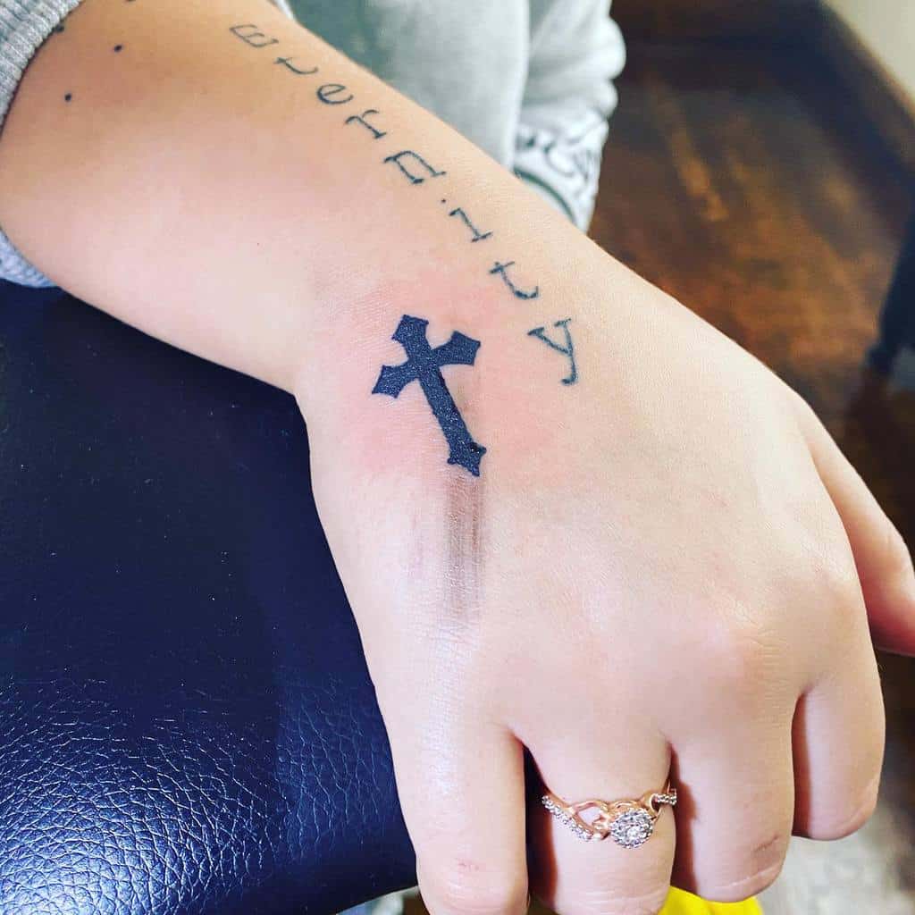 A tattooed hand displays a bold black cross alongside the word "eternity" in elegant cursive