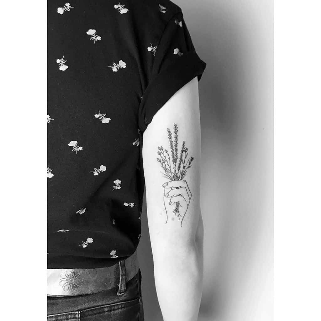 A tattoo on the upper arm depicting a delicate hand holding a bundle of lavender stems. The design is in black ink with fine line details, giving the tattoo a soft and minimalistic aesthetic.