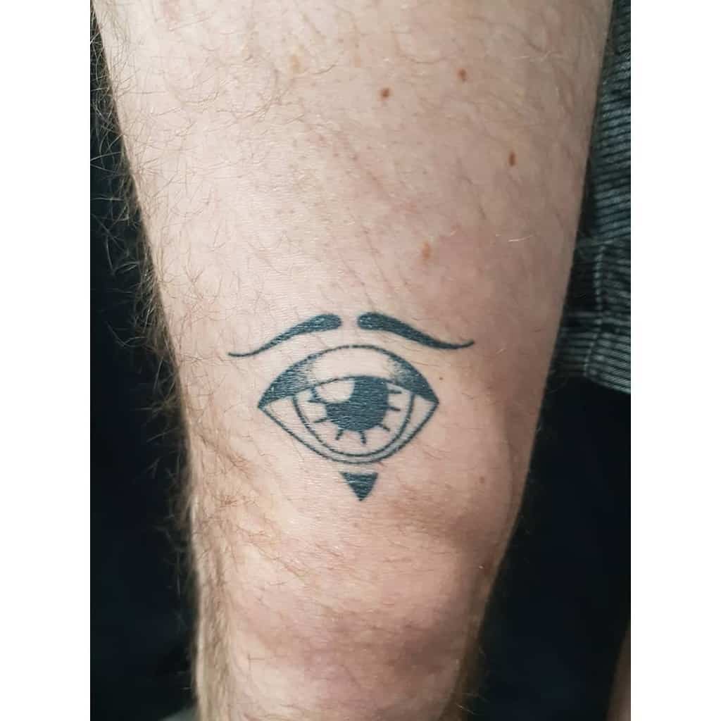 Minimalist tattoo of an eye with curved eyebrows and a small triangle below, inked in black on the leg.