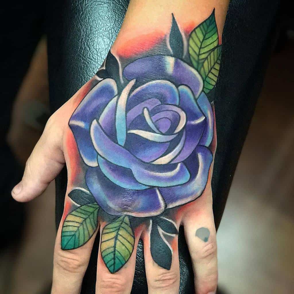 Hand tattoo features a vibrant purple rose with shades of blue, detailed green leaves, and a bold design