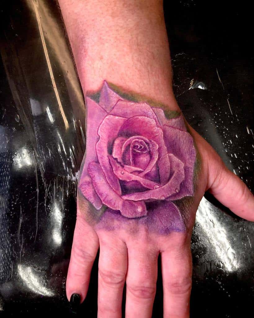 Adorning the back of the hand, a purple rose tattoo blooms elegantly, showcasing its vibrant hues and delicate design