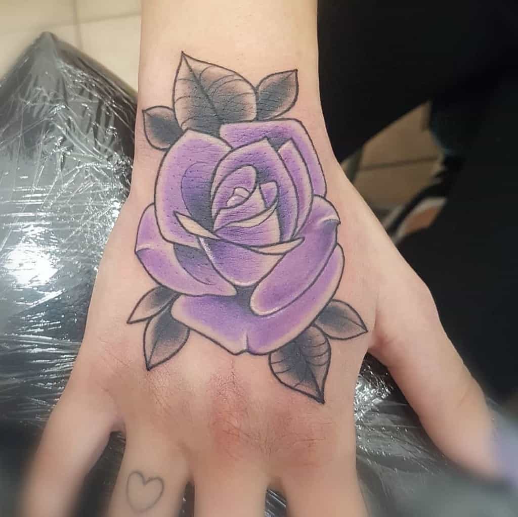 Hand tattoo of a vibrant purple rose with leaves, complemented by a small heart on the finger