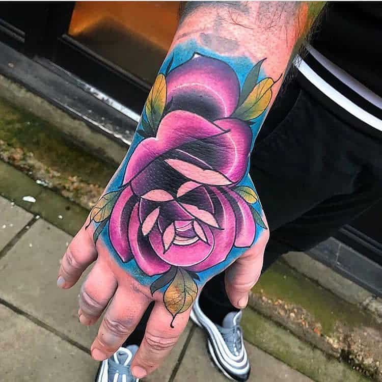 A hand tattoo of a vibrant pink, blue, and purple rose with lush green leaves