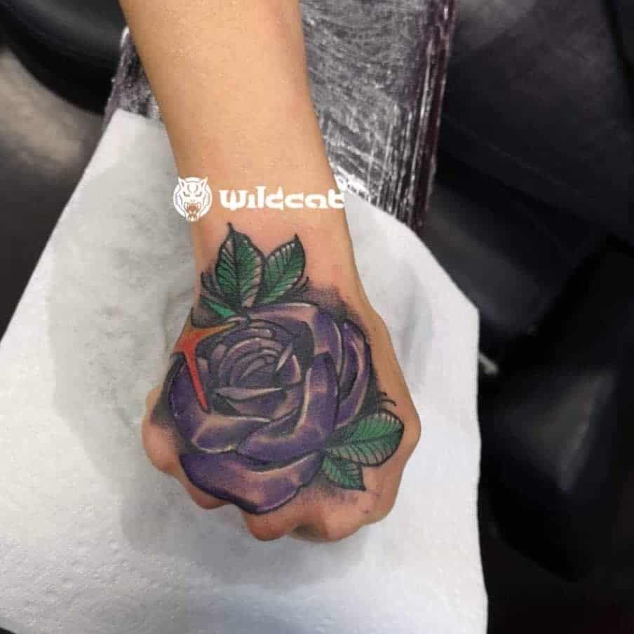 A stunning tattoo featuring a vibrant purple rose with lush green leaves elegantly graces the back of a hand