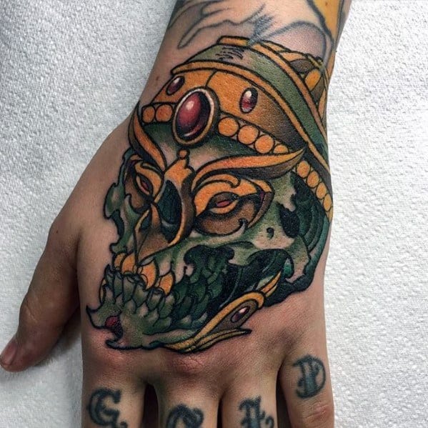 Green Tibetan skull hand tattoo with gold crown, red jewel centerpiece, and bold black outlines for a fierce and striking design.
