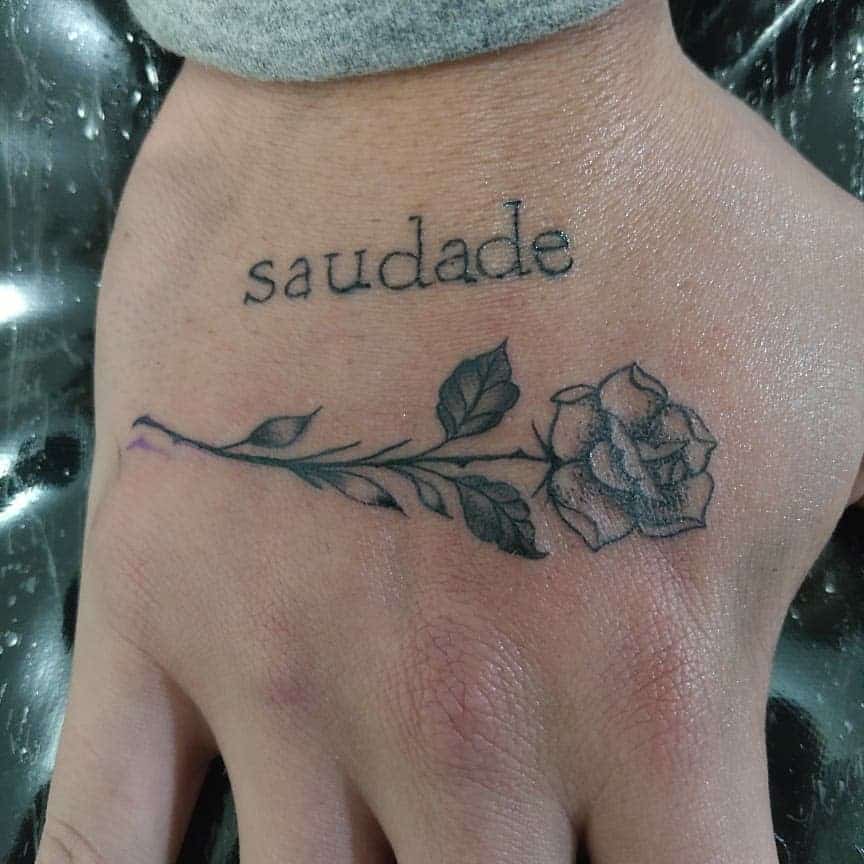 A hand proudly displays a "saudade" tattoo, elegantly adorned with a rose design below it, where the delicate stem winds gracefully down the wrist