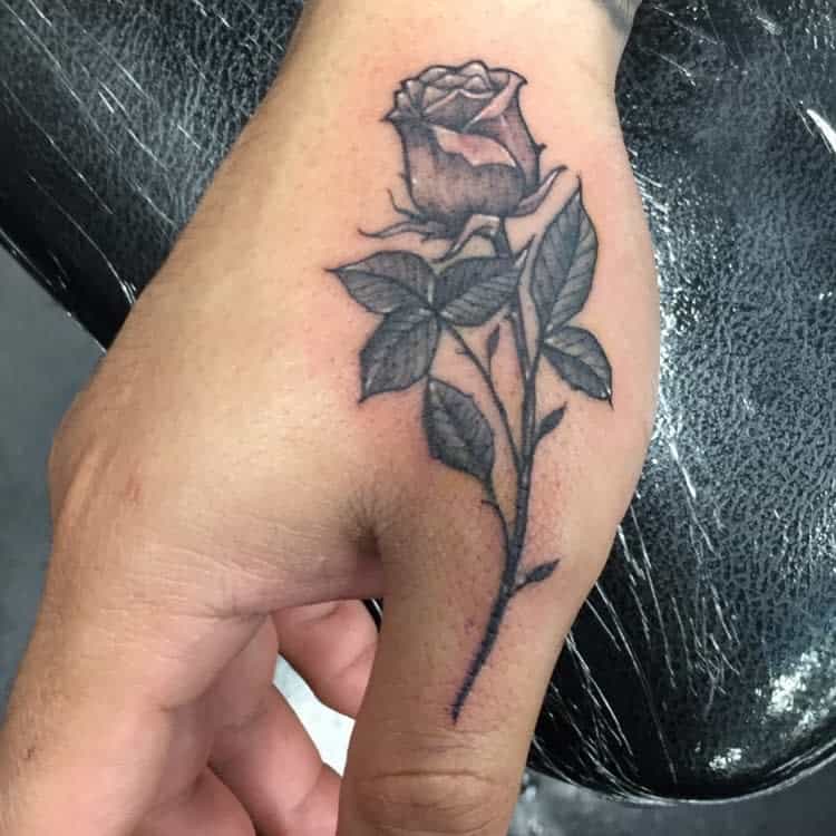 A hand tattoo features a detailed rose with leaves and a slender stem elegantly trailing down the back of the hand