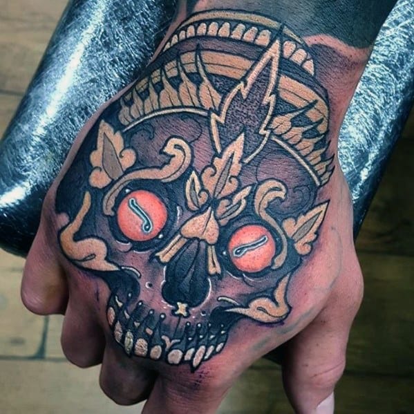 Tibetan skull tattoo on the hand with red eyes, bold black outlines, and stylized crown elements in muted gold and peach tones.