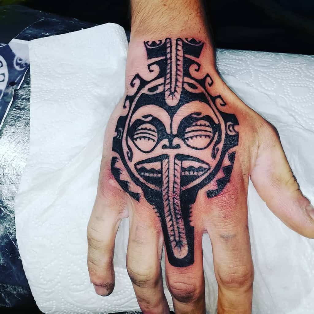 A bold black tribal tattoo of a Tiki mask, featuring intricate patterns and geometric shapes