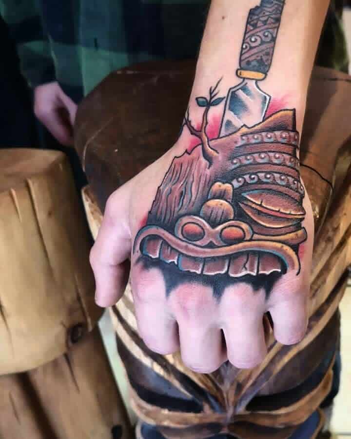 Vibrant tattoo of a Tiki mask made with wood and has leaves and branch growing,  with a knife, surrounded by tribal patterns and shading