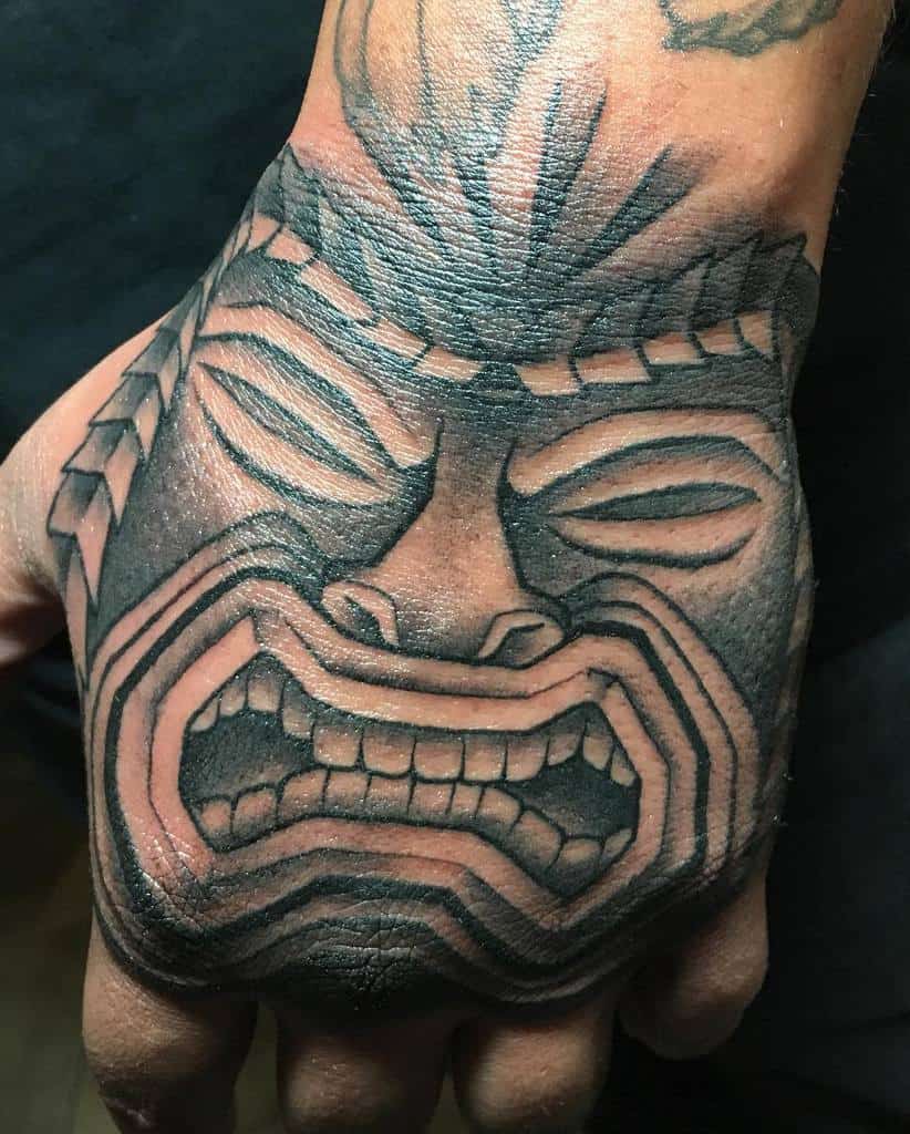 A detailed black-and-gray tattoo of a Tiki mask, with intricate lines and shading