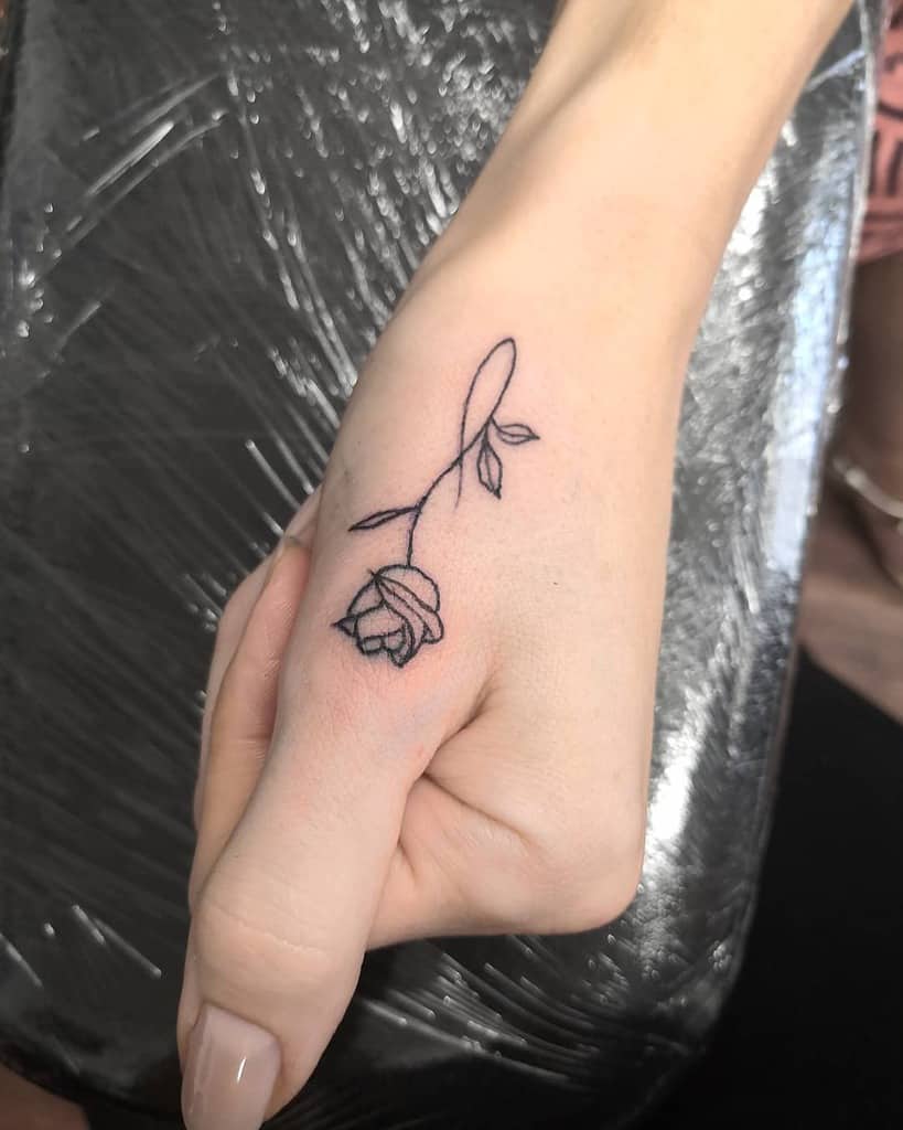 Minimalist rose tattoo on the side of a person's hand, with a long stem and several leaves