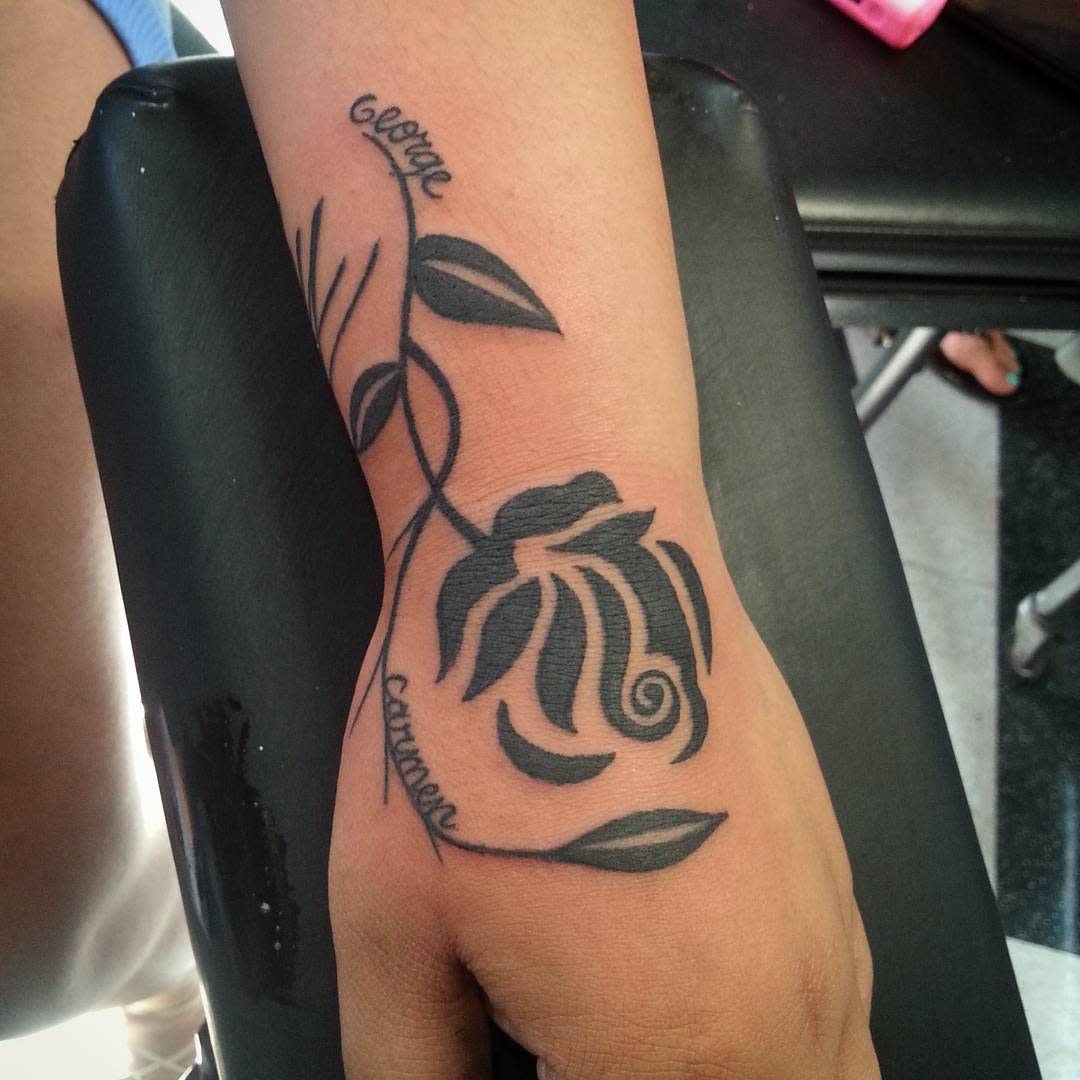 A tribal-inspired black rose tattoo elegantly adorns a person's forearm, with intertwined names woven seamlessly into its intricate design