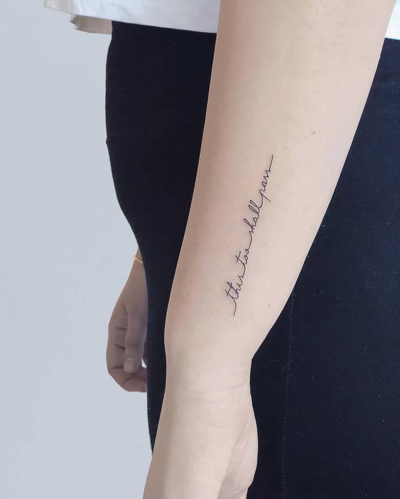 A "This Too Shall Pass" tattoo in cursive on a person's forearm, wearing a simple black outfit.