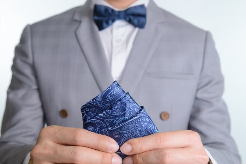 A person in a light gray suit with a blue bow tie adjusts a blue patterned pocket square