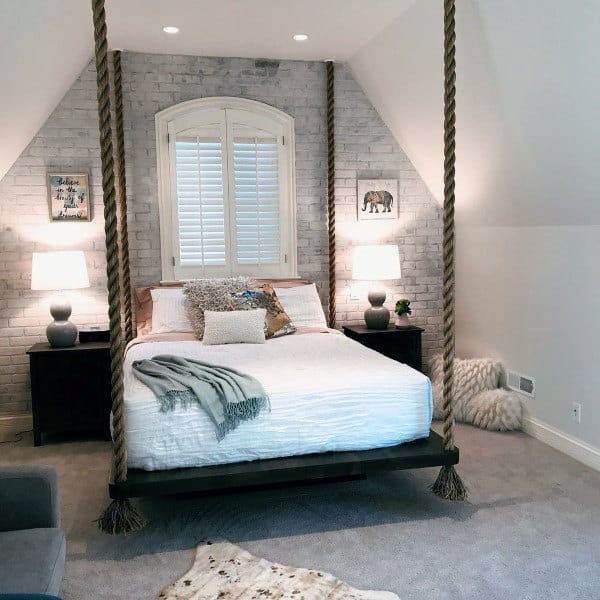 Elegant hanging bed with thick rope supports, white bedding, and a brick accent wall in a cozy room.