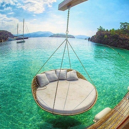 Round hanging daybed with cushions suspended over crystal-clear turquoise water.