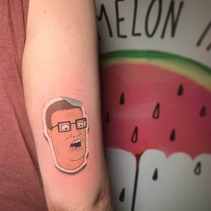 A humorous tattoo of a spoon with a man's face inside, designed in a minimalist black ink style, placed on the forearm.