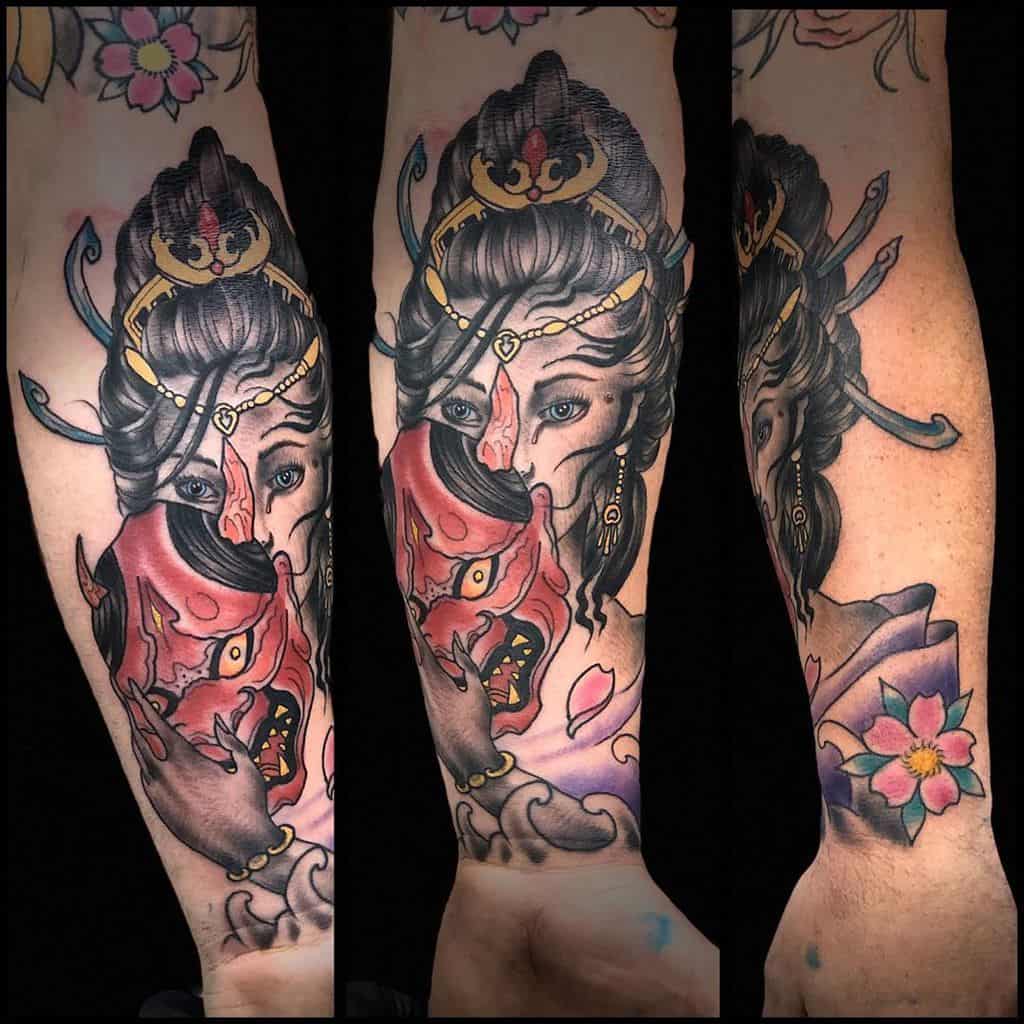 Colorful tattoo of a Geisha holding a red oni mask, revealing one eye, with intricate hair ornaments, cherry blossoms, and flowing details.