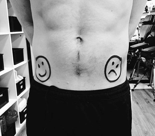 Black and white tattoo of a happy face emoji on the left hip and a sad face emoji on the right hip