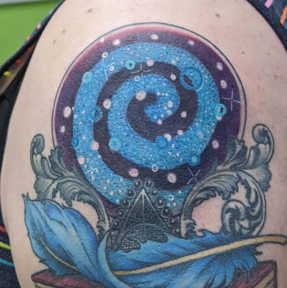 Colorful Deathly Hallows tattoo featuring a glowing blue spiral galaxy, ornate scrollwork, a quill, and a mystical crystal ball design.