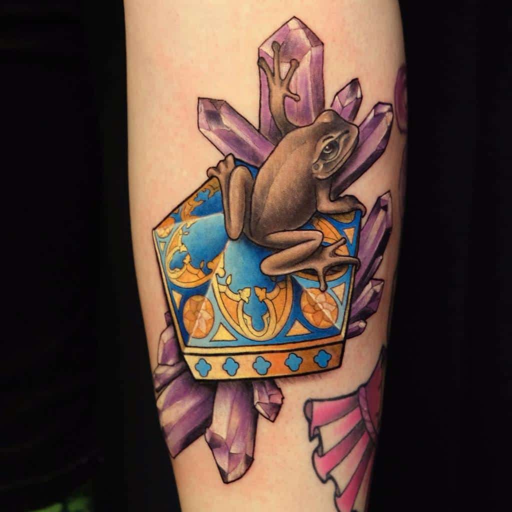 Detailed Chocolate Frog tattoo from Harry Potter, featuring its ornate blue and gold box surrounded by vibrant purple crystals