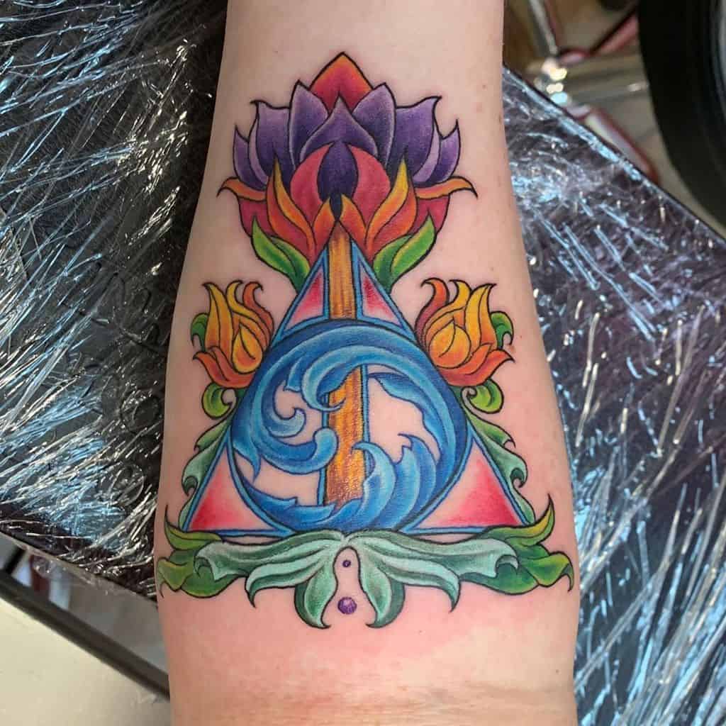 Vibrant Deathly Hallows tattoo with floral elements, featuring a lotus, swirling blue waves, and fiery accents, blending magic and nature