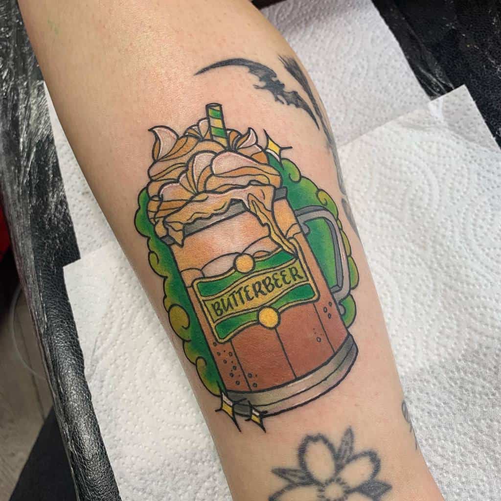 Colorful Butterbeer tattoo inspired by Harry Potter, featuring a frothy mug with whipped topping, a green decorative frame, and flying bats