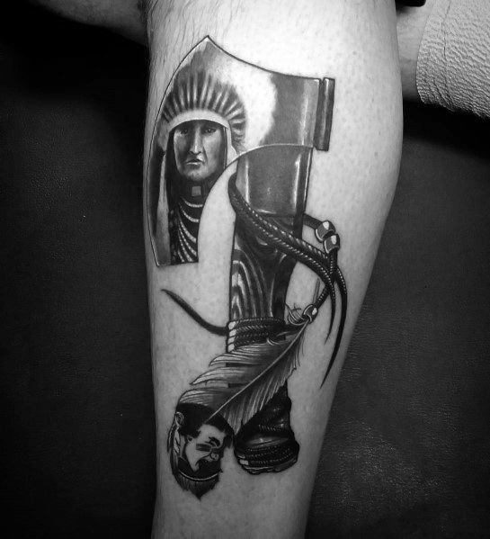 Black and grey hatchet tattoo with a detailed Native American chief's face in the blade and a realistic warrior figure wrapped around the handle.