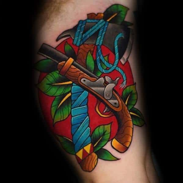 Colorful traditional tattoo featuring a hatchet and flintlock pistol crossed over a red background, with blue wraps, green leaves, and bold shading.
