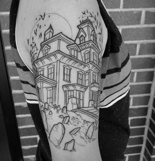 Upper arm tattoo of a haunted mansion with gravestones in front, a full moon in the background, and bats flying overhead.