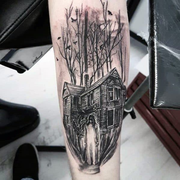 Forearm tattoo of a haunted house in the woods with a ghostly figure in front, leafless trees, and bats flying in a spooky sky.