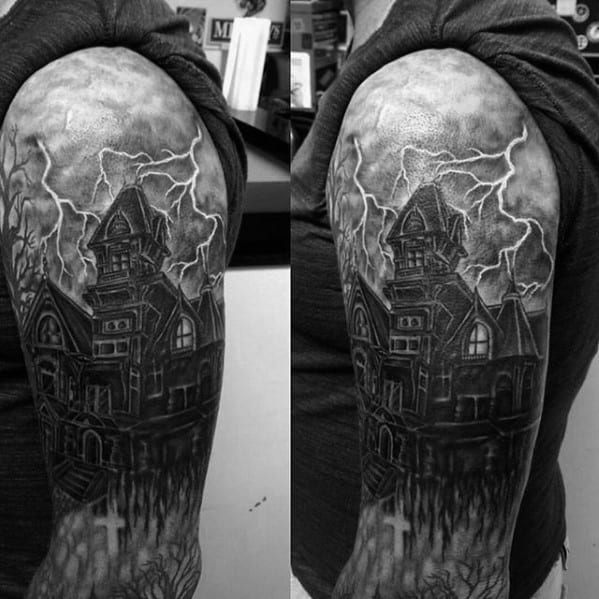 Upper arm tattoo of a haunted mansion under a stormy sky, with lightning bolts, dark clouds, and grave markers at the bottom.