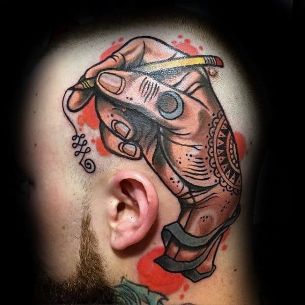 Tattoo of a hand grasping a pencil is intricately designed on a person's shaved head, enhanced with bold red shading accents