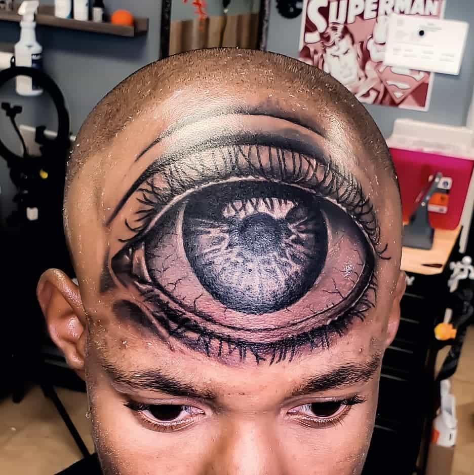 Tattoo of a highly detailed, realistic third eye inked in black and grey shading across the forehead.