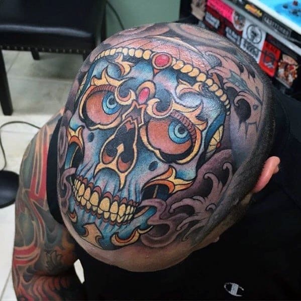 Vivid Tibetan skull head tattoo with blue eyes, golden crown, red gem, and swirling smoke details covering the entire scalp.