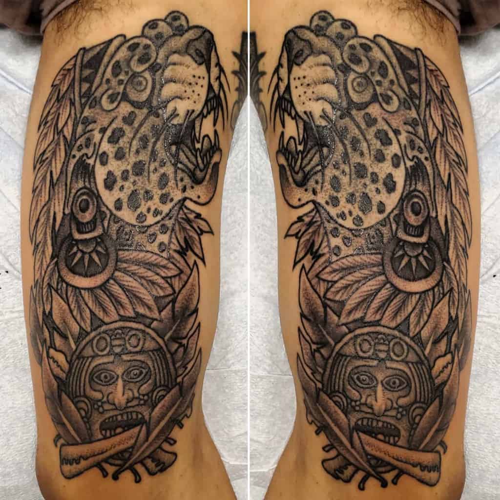 A detailed tattoo featuring a roaring leopard's head surrounded by feathers and intricate patterns, with a tribal mask at the base, blending nature and cultural elements.
