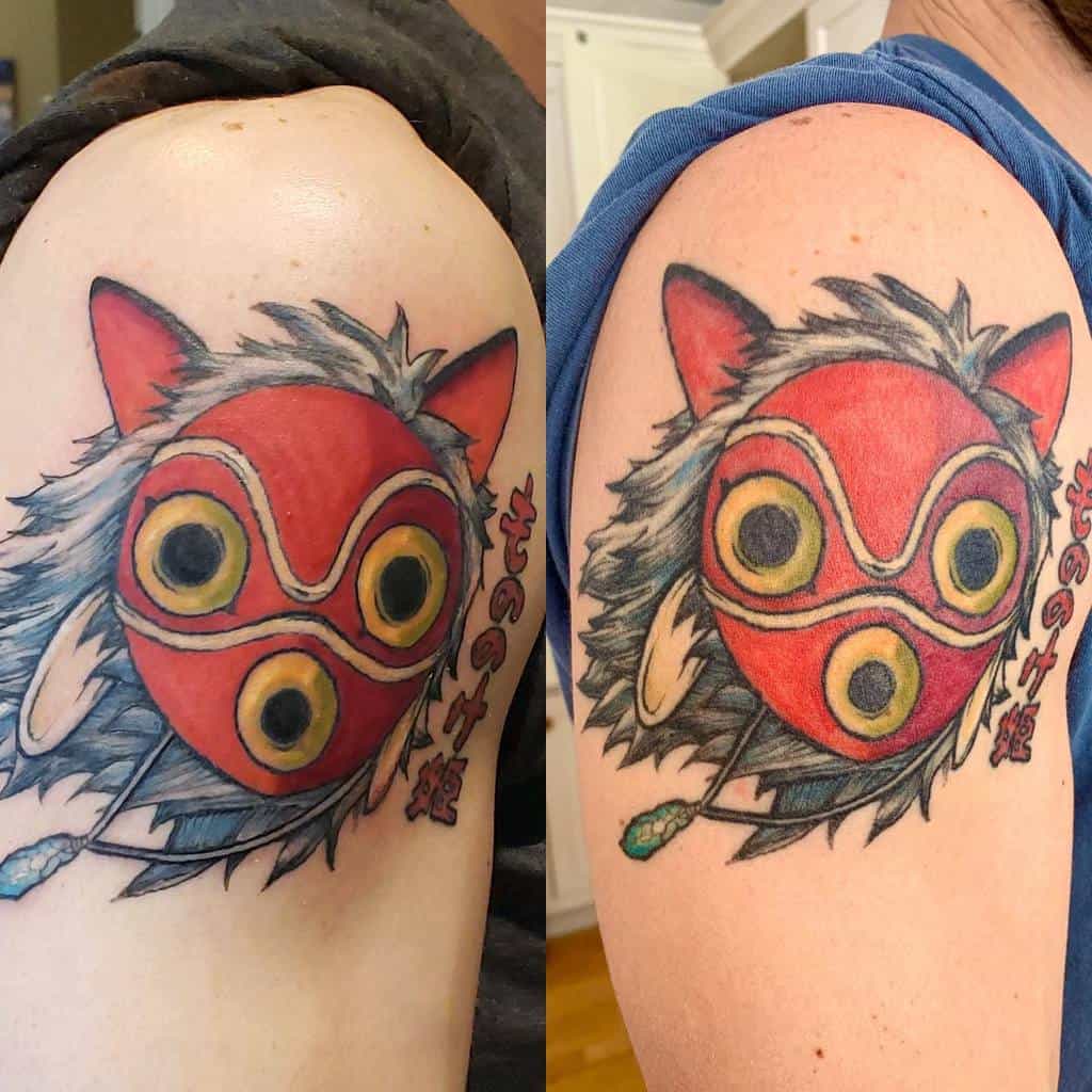 Vibrant tattoo of San’s red wolf mask, with detailed fur and feather accents, and the text 'Mononoke' in Japanese, on the upper arm.