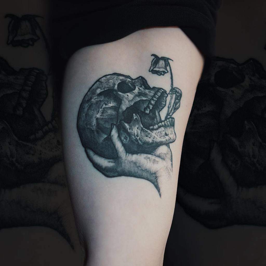 Black and grey tattoo of a skull held in a hand with a wilting rose, symbolizing mortality and transience, inked on the upper thigh.