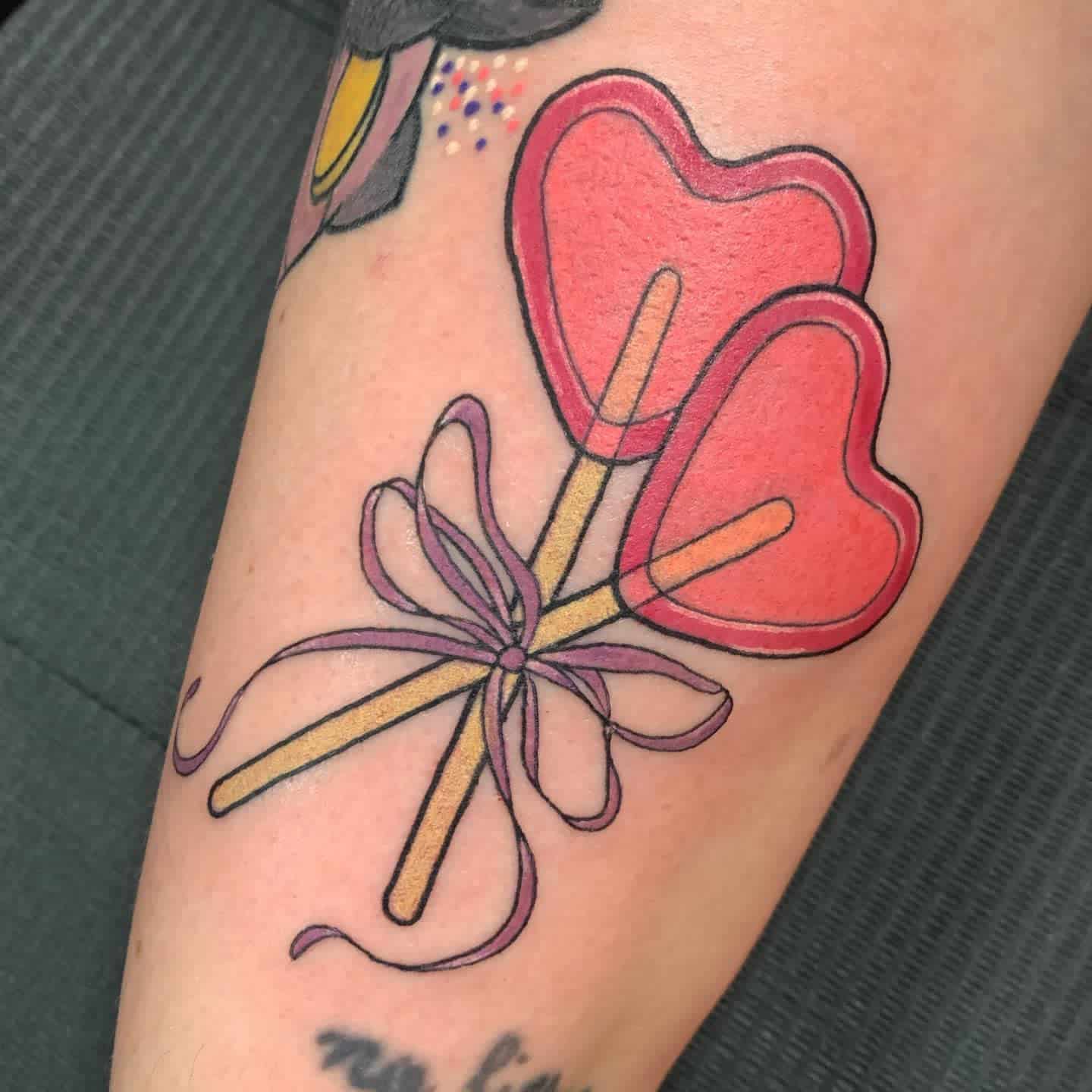 Tattoo of two red heart-shaped lollipops with sticks tied together by a purple ribbon