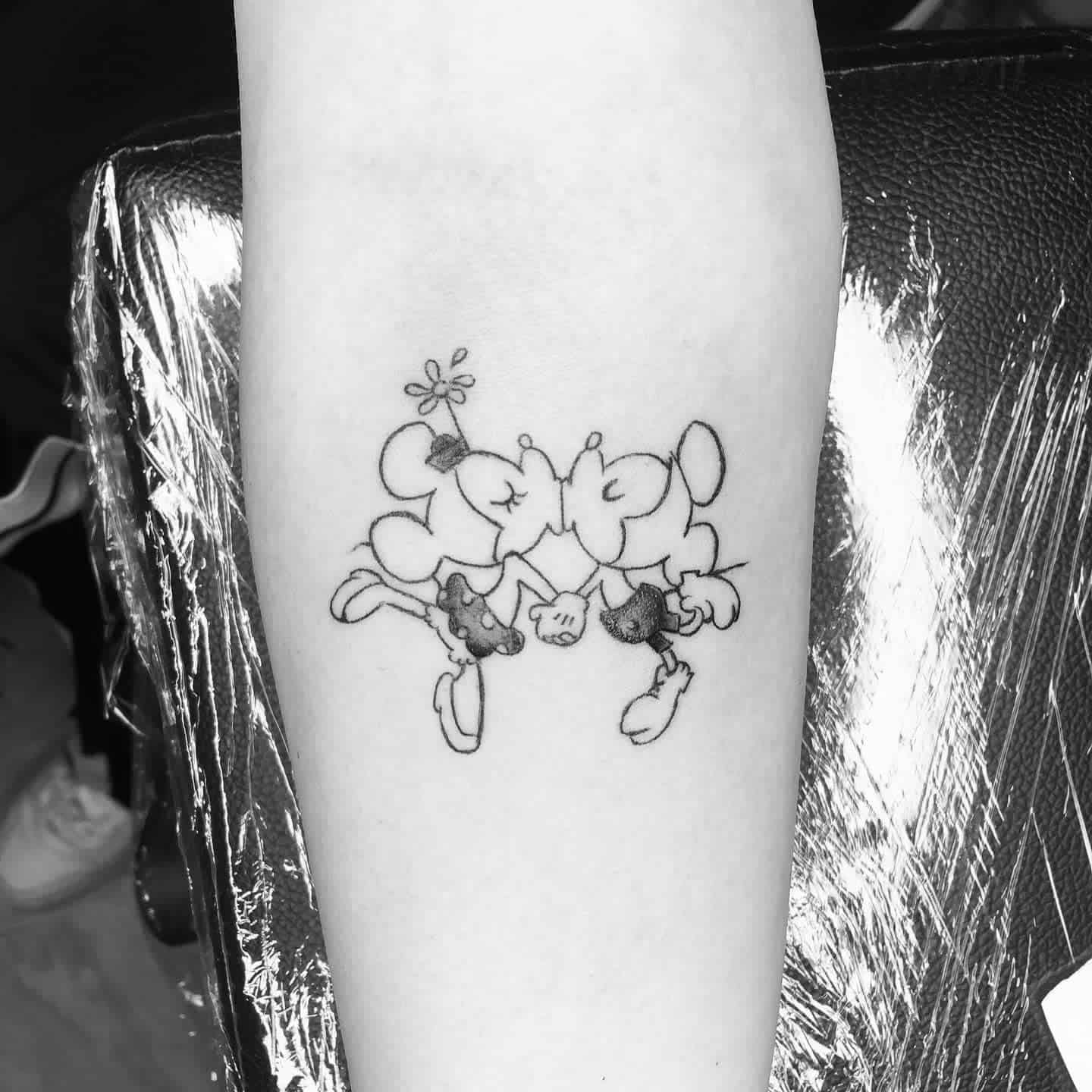 Black and white tattoo of Minnie Mouse with flower on her head and Mickey Mouse, holding hands and kissing