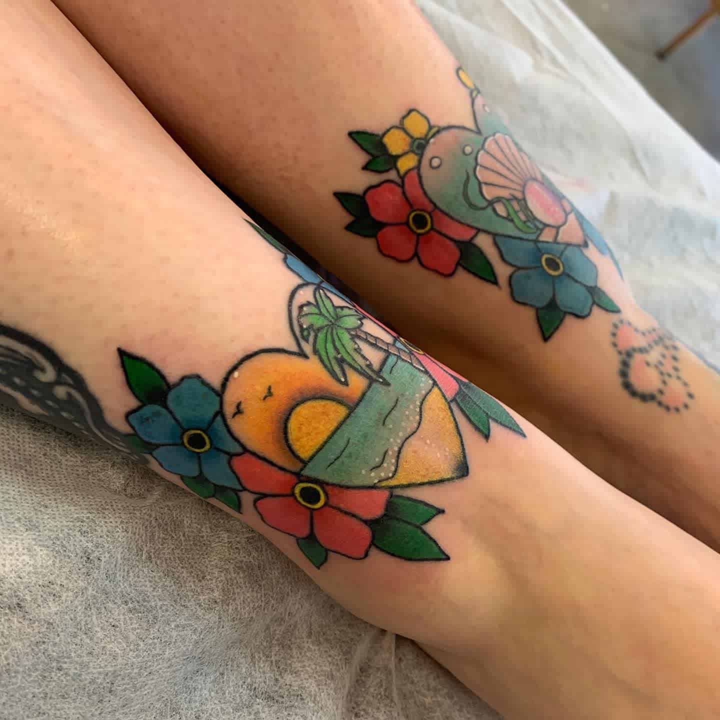 Vibrant tattoos on both calves featuring tropical scenes with hearts, a sunset, a palm tree, and shells, surrounded by colorful flowers in red, blue, and green