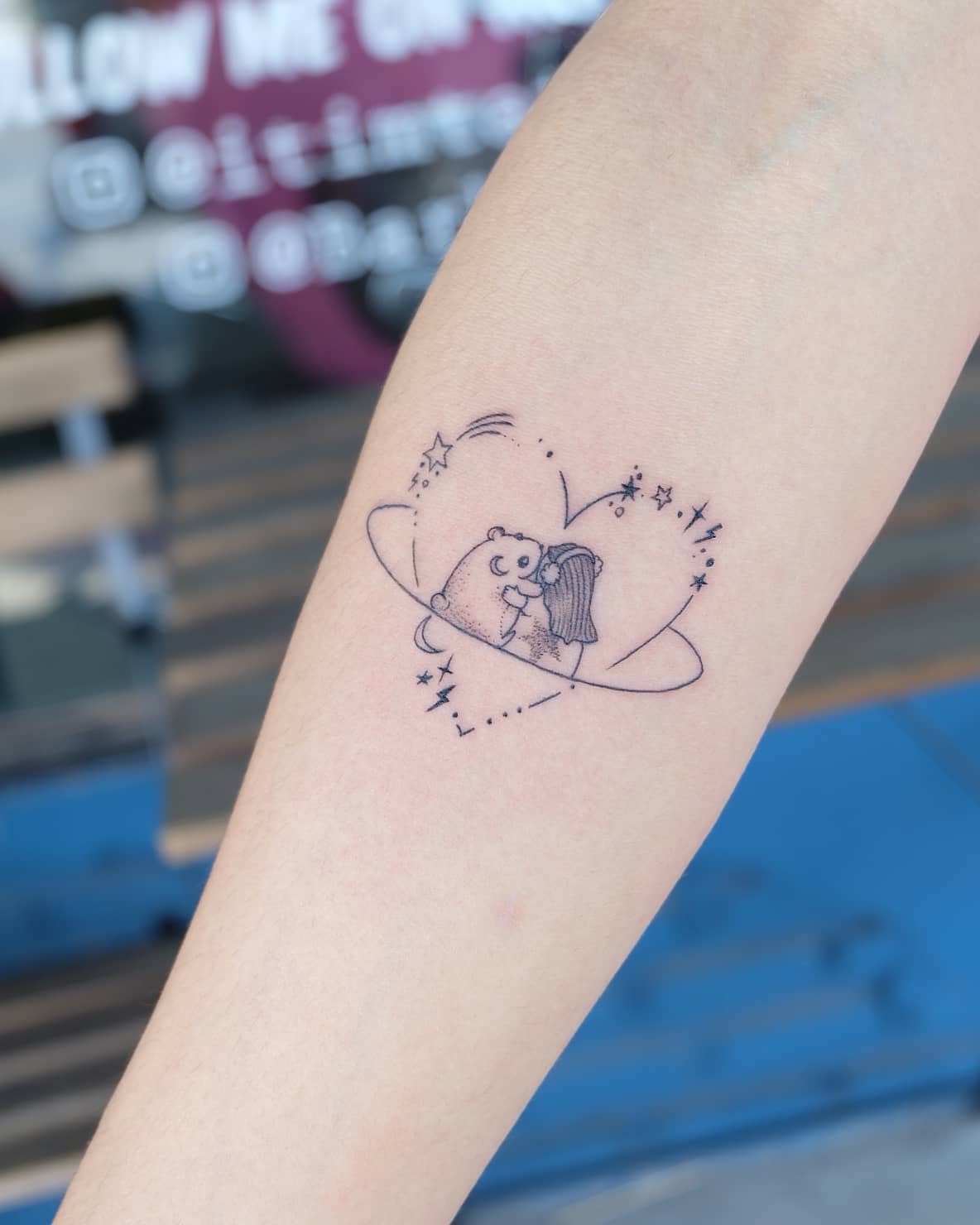 A tattoo of a bear hugging a girl, surrounded by stars and a heart-shaped orbit