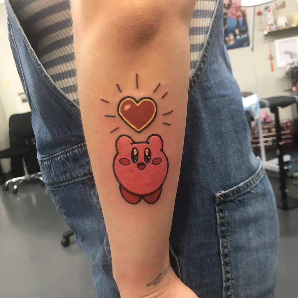 Cute Kirby tattoo on forearm with a golden-outlined red heart floating above, emitting radiant lines