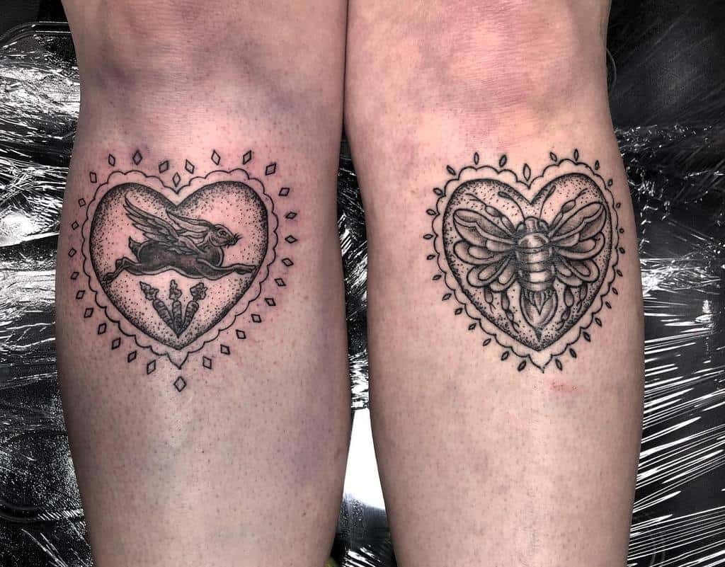 Black and gray tattoos of a rabbit with wings, three carrots below and a bee, both enclosed in heart-shaped frames with dotted patterns and ornate details