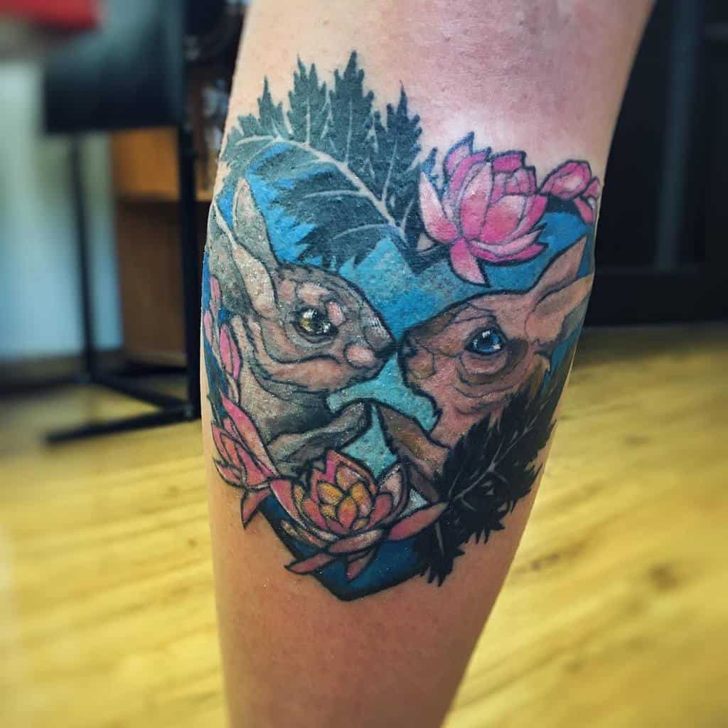 Colorful tattoo of two rabbits within a blue heart-shaped design, surrounded by lush leaves and vibrant lotus flowers